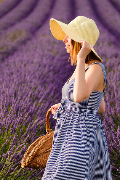 Молода леді, прикрашена солом'яним капелюхом, чудово стоїть на лавандовому полі, витончено носить плетений кошик, наповнений яскраво-фіолетовими квітами
. - Фото, зображення