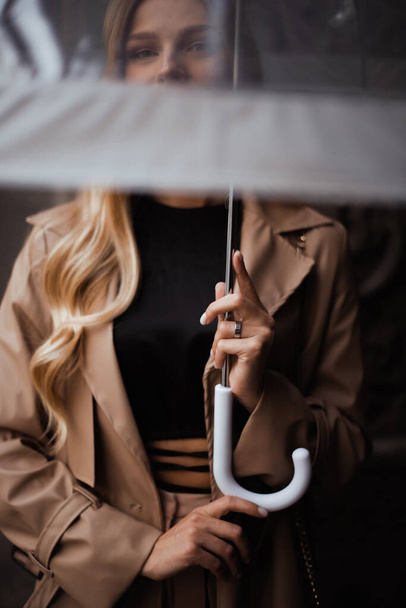 Yağmurlu günlerde şeffaf şemsiyeyi tutan ellere odaklan. Kadın kameraya şeffaf şemsiyeyle bakar. - Fotoğraf, Görsel