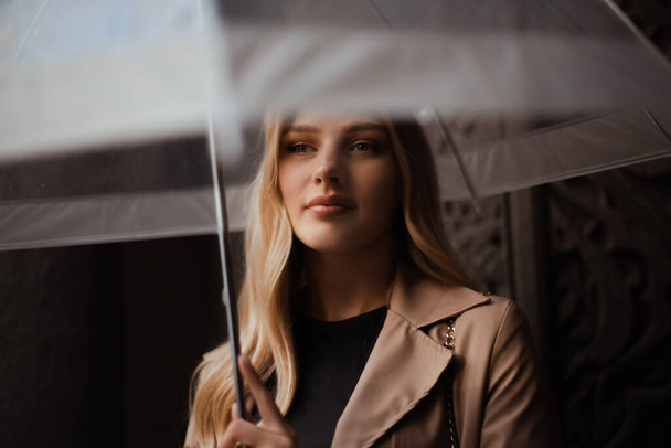 vrouw in beige jas met doorzichtige paraplu in regenachtige straat scene - Foto, afbeelding