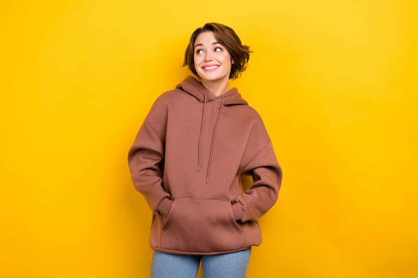 Portrait of youngster lady look side empty space good mood hold hands pockets hood shirt isolated yellow color background. - Photo, image