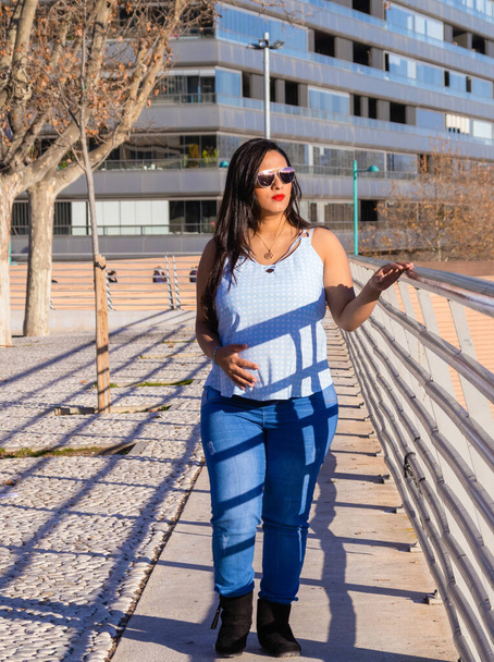 Young pregnant woman walking by a fence in the city, looking at the horizon while touching her belly, wearing a blue blouse, jeans and sunglasses. - Foto, Bild