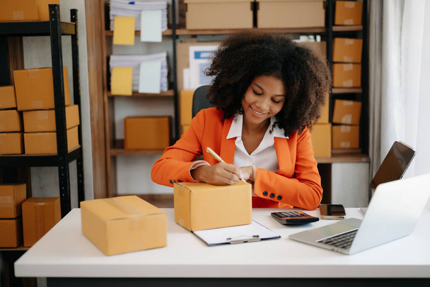 Startup small business SME, Entrepreneur owner African woman using smartphone and laptop taking receive and checking online purchase shopping order to prepare pack product boxes at home office - Foto, afbeelding