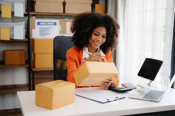 Startup small business SME, Entrepreneur owner African woman using smartphone and laptop taking receive and checking online purchase shopping order to prepare pack product boxes at home office - 写真・画像