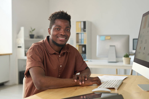 Muotokuva Afrikkalainen amerikkalainen nuori liikemies hymyilee kameralle istuessaan työpaikallaan - Valokuva, kuva