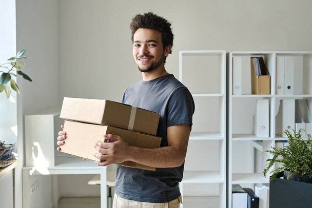 Portrait of young man smiling at camera while carrying packed boxes in office - Φωτογραφία, εικόνα