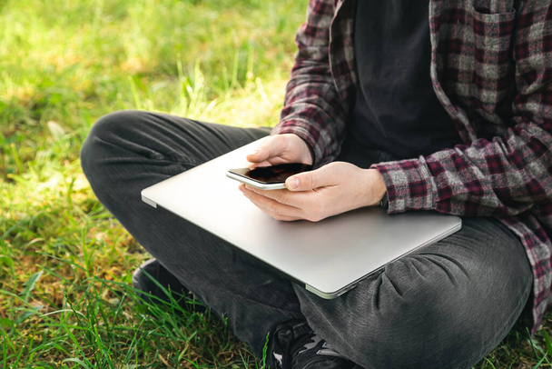 屋外のガジェットを使用して、彼の手にノートパソコンやスマートフォンで芝生の上に座っている男、クローズアップ. - 写真・画像