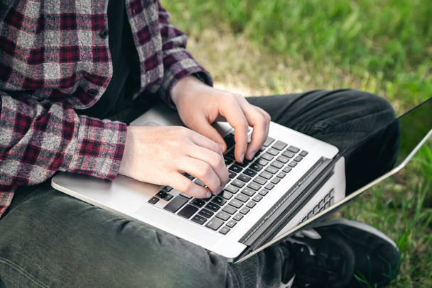 A man in the park sitting on the grass with a laptop, close-up hands and laptop, outdoor work in summer. - Фото, изображение