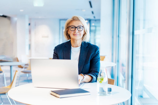 Portrait of 50s confident mature businesswoman looking at camera, middle-aged experienced senior female professional working on laptop in open space office. Female entrepreneur working remotely. - 写真・画像