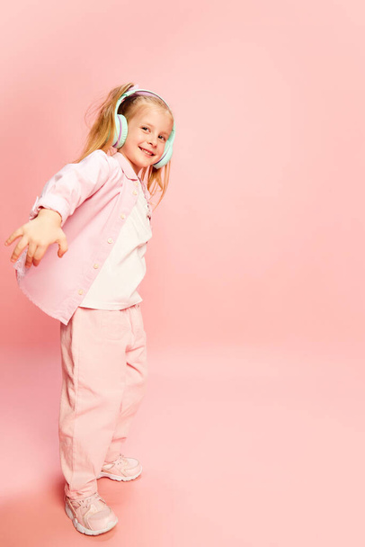Full-length portrait of happy, beautiful little girl, child listening to music in headphones, smiling and dancing against pink background. Concept of childhood, emotions, lifestyle, fashion, joy. Ad - Фото, зображення