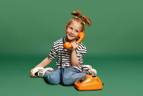 Portrait of little girl, child with cute, funny hairstyle, playing, sitting on floor and talking on retro phone against green studio background. Concept of childhood, emotions, lifestyle, joy. Ad - Foto, Bild
