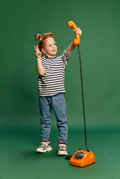 Портрет маленькой девочки, ребенка с милой, смешной прической, играющей, делающей селфи с ретро-телефоном на зеленом фоне студии. Концепция детства, эмоций, образа жизни, моды, радости. Объявление - Фото, изображение