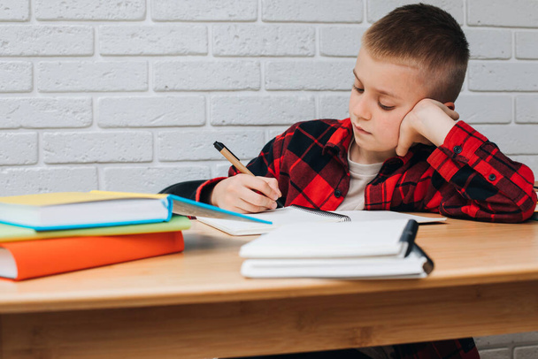 Wracajcie do szkoły. Chłopiec w trakcie nauki siedzi przy stole z książkami, notatnikami i ołówkami. Widok z przodu - Zdjęcie, obraz