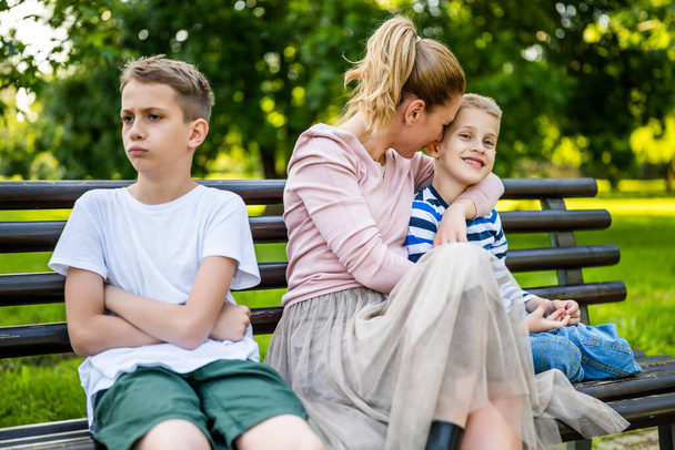 Счастливая мать сидит со своими сыновьями на скамейке в парке. Один мальчик обижен и дуется.. - Фото, изображение