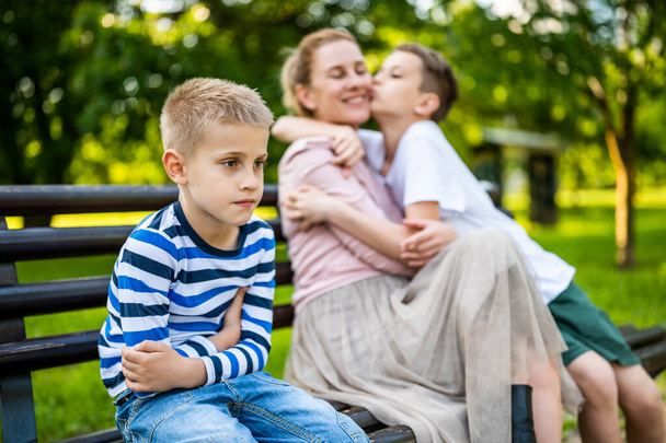 Feliz madre está sentada con sus hijos en el banco en el parque. Un chico se ofende y hace pucheros.. - Foto, Imagen