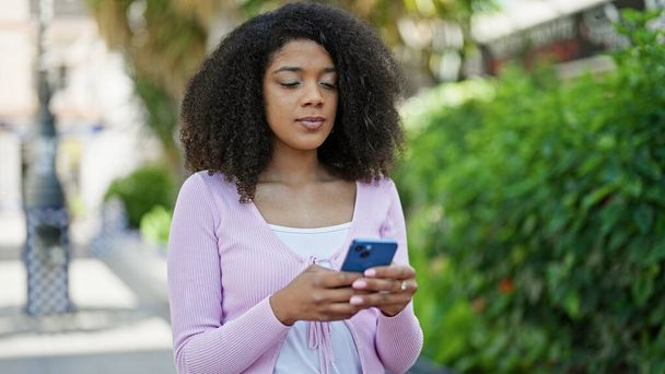 Африканська американка, яка користується смартфоном з серйозним виразом обличчя в парку. - Фото, зображення