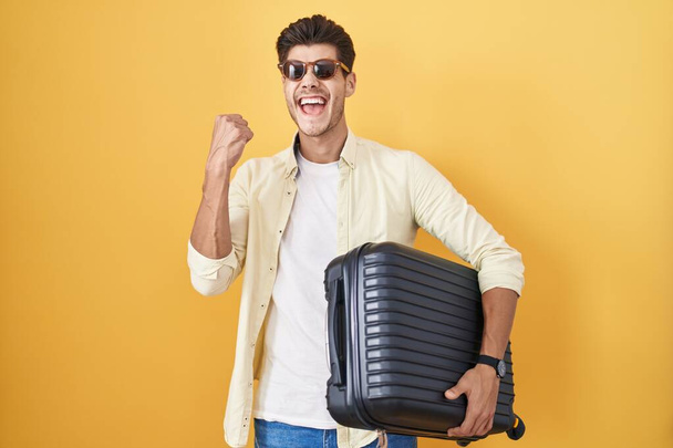 Yaz tatilinde valiz tutan genç İspanyol adam çok mutlu ve heyecanlı bir şekilde kolları havada, gülümseyen ve başarı çığlıkları atan bir jest yapıyor. kutlama konsepti.  - Fotoğraf, Görsel