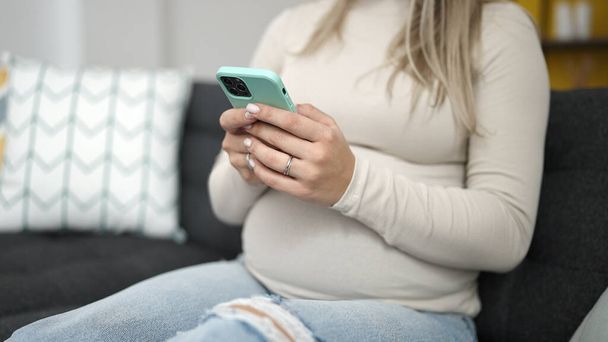 Mujer embarazada joven usando teléfono inteligente sentado en el sofá en casa - Foto, Imagen