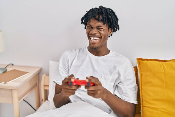 Αφροαμερικάνος που παίζει βιντεοπαιχνίδι καθισμένος στο κρεβάτι στην κρεβατοκάμαρα - Φωτογραφία, εικόνα