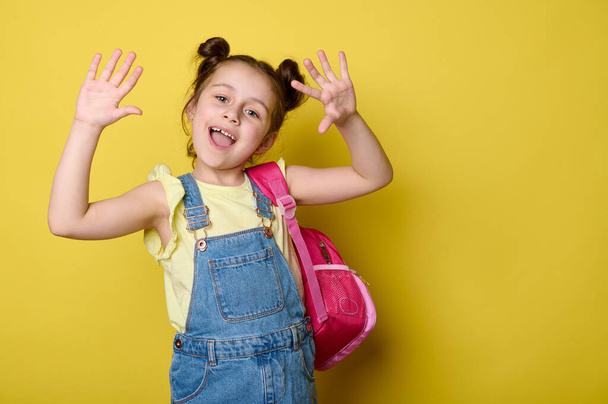 Veselý školačka dívka ukazuje na kameru její ruce dlaně nahoru, usmívá se, těší se na začátku nového školního roku, pózovat s růžovým batohem, oblečený v modrých džínových šatech, izolované přes žluté pozadí - Fotografie, Obrázek