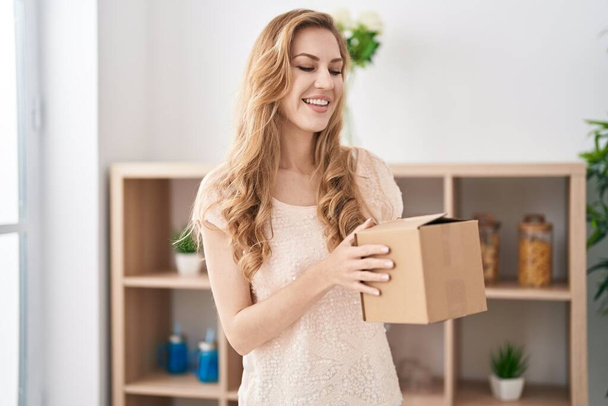 Joven mujer rubia sonriendo confiado sosteniendo paquete en casa - Foto, imagen