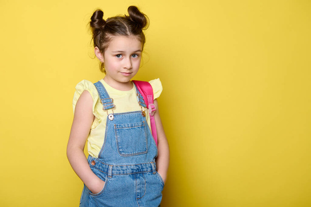 Retrato de estúdio no fundo amarelo de uma menina linda caucasiana 6 anos em macacão jeans azul, carregando uma mochila, segurando as mãos nos bolsos e olhando com confiança para a câmera. Espaço publicitário cópia - Foto, Imagem