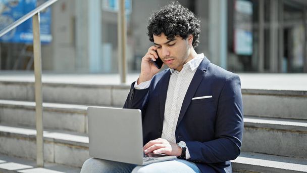 Nuori latinomies liike-elämän työntekijä käyttää kannettavaa tietokonetta puhumalla älypuhelimella kadulla - Valokuva, kuva