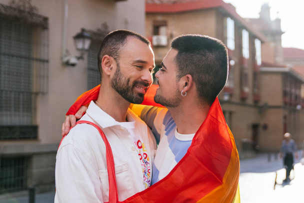 In een zonovergoten stad, twee homo vrienden houden elkaar vast, uitwisselen een tedere blik, versierd met levendige LGBT symbolen. - Foto, afbeelding