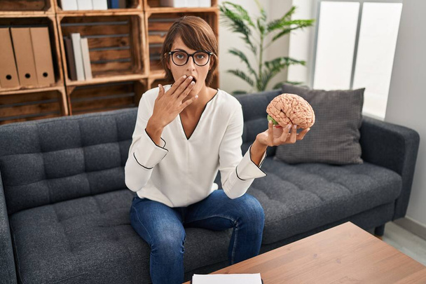 Brunette woman working at therapy office holding brain covering mouth with hand, shocked and afraid for mistake. surprised expression  - Zdjęcie, obraz