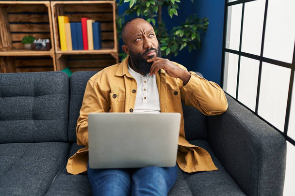 African american man using laptop at home sitting on the sofa serious face thinking about question with hand on chin, thoughtful about confusing idea  - Fotografie, Obrázek