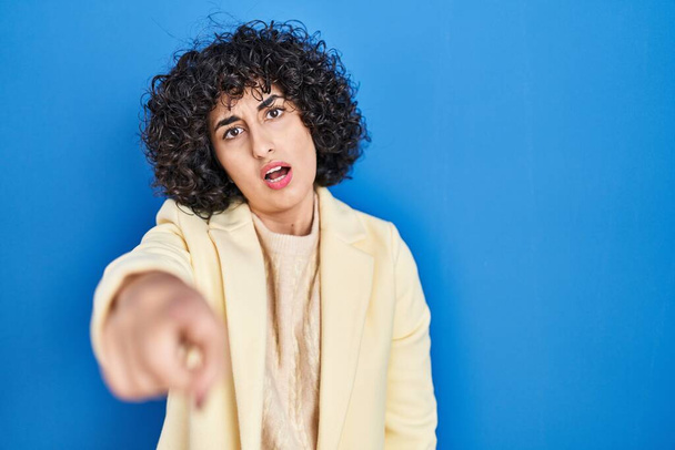 Jeune femme brune avec les cheveux bouclés debout sur fond bleu pointant mécontent et frustré à la caméra, en colère et furieux contre vous  - Photo, image