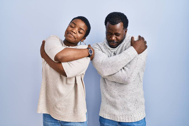 Young african american couple standing over blue background together hugging oneself happy and positive, smiling confident. self love and self care  - Foto, Imagem