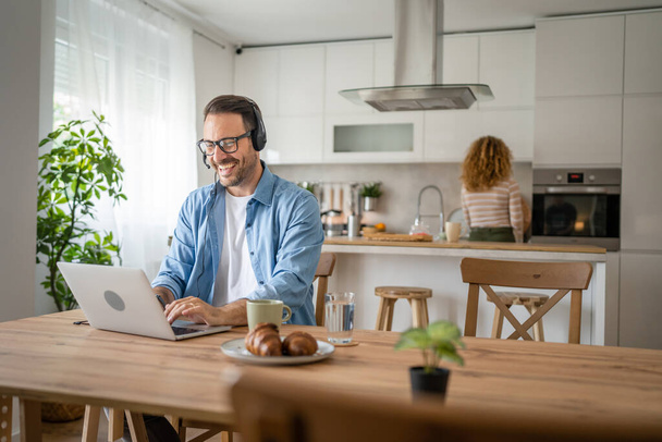 Een man Kaukasische man freelancer werken vanuit huis met koptelefoon op zijn hoofd op laptop computer customer support gelukkig glimlach vertrouwen zijn vrouw staan op de achtergrond - Foto, afbeelding