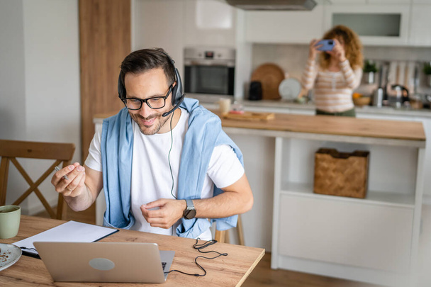 Ein Mann kaukasischen männlichen Freiberufler arbeiten von zu Hause aus mit Kopfhörern auf dem Kopf auf Laptop-Computer Kundenbetreuung glückliches Lächeln zuversichtlich, dass seine Frau im Hintergrund stehen - Foto, Bild
