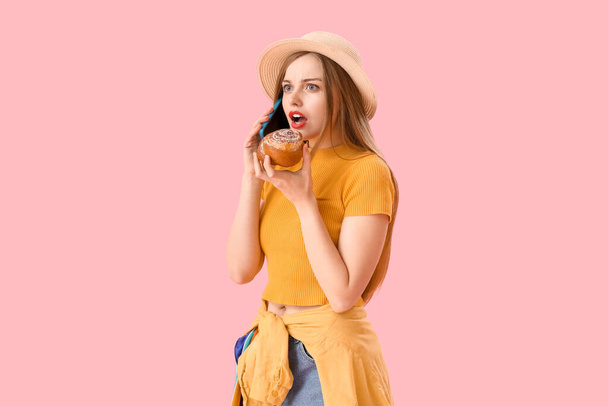 Shocked young woman with tasty bun talking by mobile phone on pink background - Photo, Image