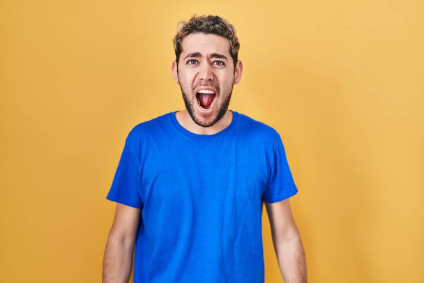 Hispanischer Mann mit Bart, der vor gelbem Hintergrund steht, wütend und wahnsinnig schreiend frustriert und wütend, schreiend vor Wut. Wut und aggressives Konzept.  - Foto, Bild