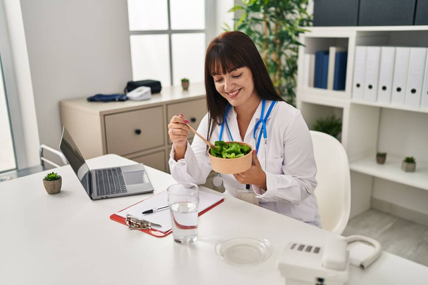 Молода красива іспаномовна жінка-лікар, використовуючи ноутбук, що їсть салат у клініці
 - Фото, зображення