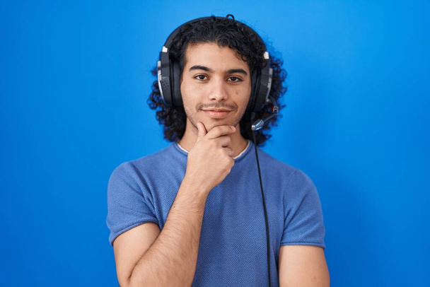 Hombre hispano con el pelo rizado escuchando música usando auriculares que miran confiados a la cámara con sonrisa con los brazos cruzados y la mano levantada en la barbilla. pensar positivamente.  - Foto, imagen