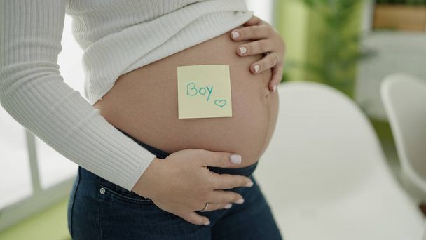 Young pregnant woman putting boy reminder paper on belly at dinning room - Foto, Imagem