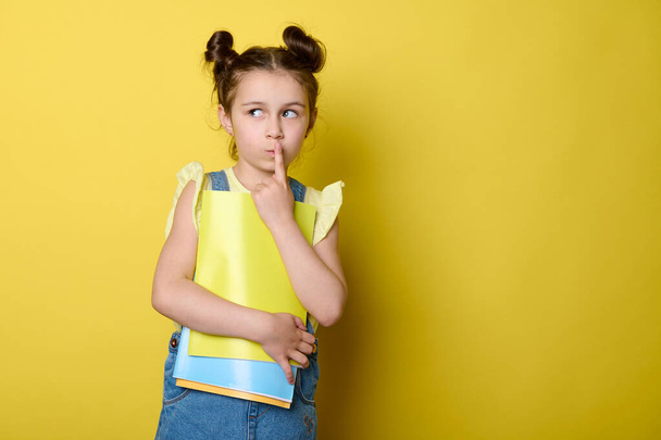 Задумчивая мечтательная школьница, прикладывающая палец к губам, рассуждающая, отводящая в сторону рекламное пространство, позирующая с красочными учебниками на желтом фоне, одетая в повседневную джинсу - Фото, изображение