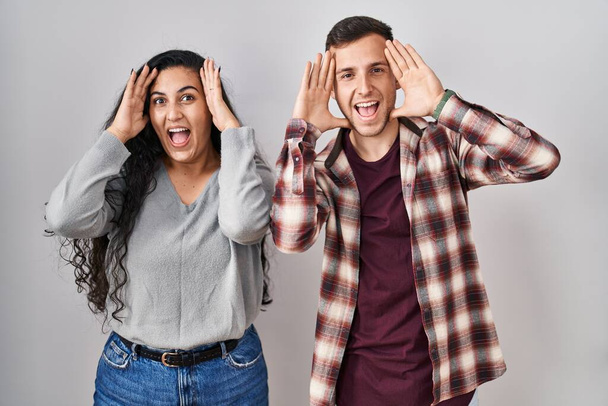 Young hispanic couple standing over white background smiling cheerful playing peek a boo with hands showing face. surprised and exited  - Fotoğraf, Görsel