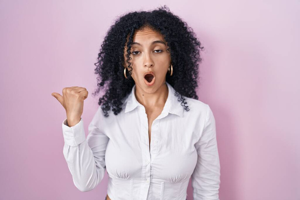 Hispanic woman with curly hair standing over pink background surprised pointing with hand finger to the side, open mouth amazed expression.  - Fotografie, Obrázek