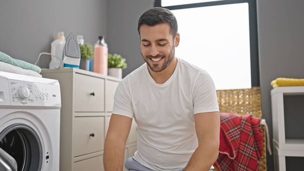 Young hispanic man smiling confident sitting on floor at laundry room - Foto, immagini