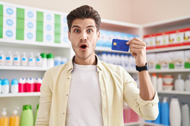 Joven hombre hispano en farmacia con tarjeta de crédito asustado y sorprendido con la boca abierta para sorpresa, cara de incredulidad  - Foto, Imagen