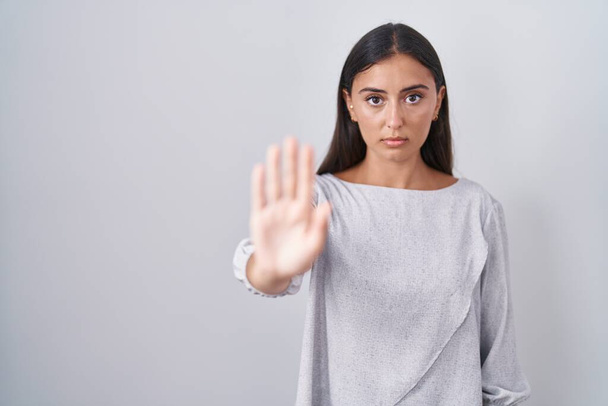 Jonge Spaanse vrouw die over een witte achtergrond staat te zingen stop met handpalm van de hand. waarschuwingsuitdrukking met negatief en ernstig gebaar op het gezicht.  - Foto, afbeelding