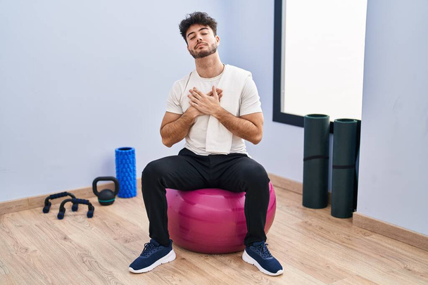 Ein spanischer Mann mit Bart sitzt auf Pilatebällen im Yoga-Raum und lächelt mit geschlossenen Augen und dankbarer Geste auf dem Gesicht. Gesundheitskonzept.  - Foto, Bild