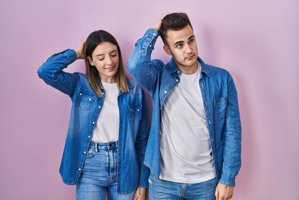 Young hispanic couple standing over pink background confuse and wondering about question. uncertain with doubt, thinking with hand on head. pensive concept.  - Photo, Image