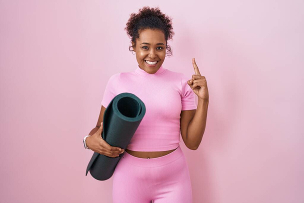 Mulher hispânica jovem com cabelo encaracolado segurando tapete de ioga sobre fundo rosa sorrindo espantado e surpreso e apontando para cima com os dedos e braços levantados.  - Foto, Imagem