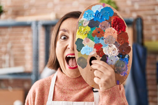 Middle age hispanic woman holding painter palette close to face celebrating crazy and amazed for success with open eyes screaming excited.  - Fotografie, Obrázek