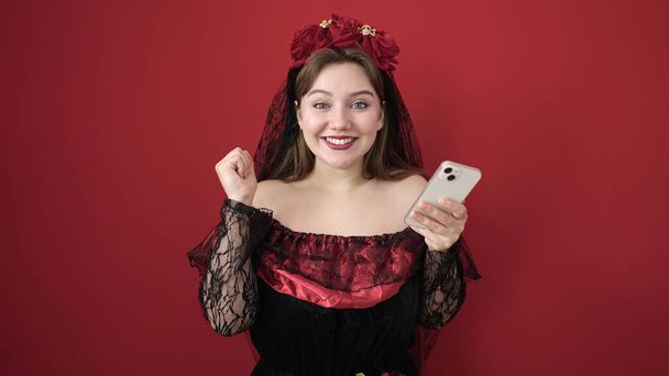 Jovem loira vestindo traje katrina usando smartphone sobre fundo vermelho isolado - Foto, Imagem