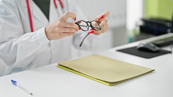 Νεαρή ξανθιά γυναίκα γιατρός χαμογελά αυτοπεποίθηση φορώντας γυαλιά στην κλινική - Πλάνα, βίντεο
